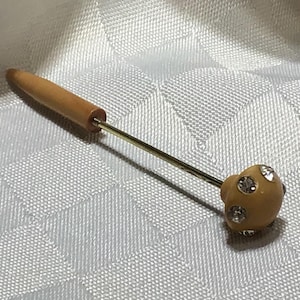 Peut inclure: Une épingle à chapeau dotée d'un long manche en bois brun clair effilé, relié à une fine tige en métal doré. L'extrémité de l'épingle se termine par un embout décoratif arrondi de couleur brun clair, incrusté de plusieurs strass transparents.
