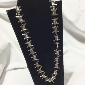 May include: A silver-coloured necklace featuring a barbed wire design. The necklace is presented on a black velvet stand against a white backdrop. The necklace is made of metal and has a repeating pattern of barbs.