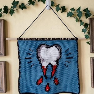 Può includere: Un arazzo da parete all'uncinetto blu con un disegno di dente con gocce di sangue rosse. Il dente è bianco con un contorno nero. L'arazzo è sospeso da un tassello di legno con una corda nera.
