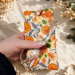 Könnte beinhalten: Ein Brillenetui mit Blumenmuster und einer Brille mit transparentem Rahmen. Das Etui hat einen cremefarbenen Hintergrund mit orangefarbenen und blauen Blumenmustern. Ein Knopf aus Perlen verschließt das Etui. Die Brille befindet sich im Etui.