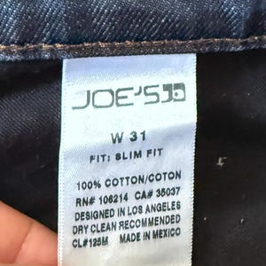 May include: A white fabric clothing tag on dark blue denim jeans. The tag reads JOE'S, W 31, FIT: SLIM FIT, 100% COTTON/COTON, RN# 106214, CA# 35037, DESIGNED IN LOS ANGELES, DRY CLEAN RECOMMENDED, CL# 125M, and MADE IN MEXICO in black text.