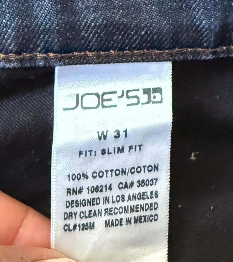 May include: A white fabric clothing tag on dark blue denim jeans. The tag reads JOE'S, W 31, FIT: SLIM FIT, 100% COTTON/COTON, RN# 106214, CA# 35037, DESIGNED IN LOS ANGELES, DRY CLEAN RECOMMENDED, CL# 125M, and MADE IN MEXICO in black text.