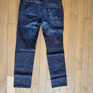 May include: A pair of dark blue denim jeans featuring a classic five-pocket design, a button fly, and subtle whiskering patterns. The back pockets show light distressing and fading, with a small rectangular brand tag on the right pocket.