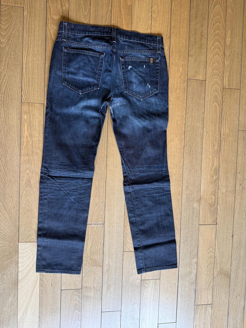 May include: A pair of dark blue denim jeans featuring a classic five-pocket design, a button fly, and subtle whiskering patterns. The back pockets show light distressing and fading, with a small rectangular brand tag on the right pocket.