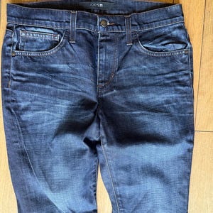May include: Dark blue denim jeans featuring a faded wash, a button and zipper fly closure, five pockets, and a waistband with belt loops. The interior label at the back of the waistband displays the brand name Joe's Jeans in white text.