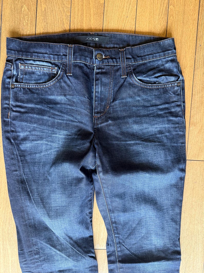 May include: Dark blue denim jeans featuring a faded wash, a button and zipper fly closure, five pockets, and a waistband with belt loops. The interior label at the back of the waistband displays the brand name Joe's Jeans in white text.