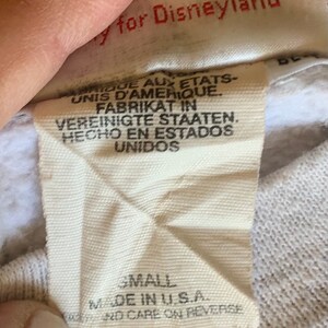 May include: White tag with black text that reads "Produced for Disneyland" and "Small Made in U.S.A. and care on reverse". The tag also has text in French, German, and Spanish.