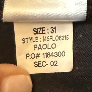 May include: A white clothing care label with black text reading SIZE: 31, STYLE: 145PL08215, PAOLO, P.O# 1184300, and SEC- 02. The top of the tag features five standard black laundry care symbols for washing, bleaching, drying, ironing, and dry cleaning.