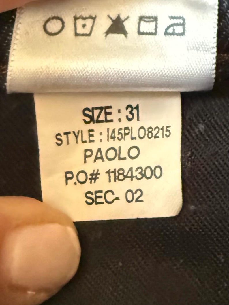 May include: A white clothing care label with black text reading SIZE: 31, STYLE: 145PL08215, PAOLO, P.O# 1184300, and SEC- 02. The top of the tag features five standard black laundry care symbols for washing, bleaching, drying, ironing, and dry cleaning.