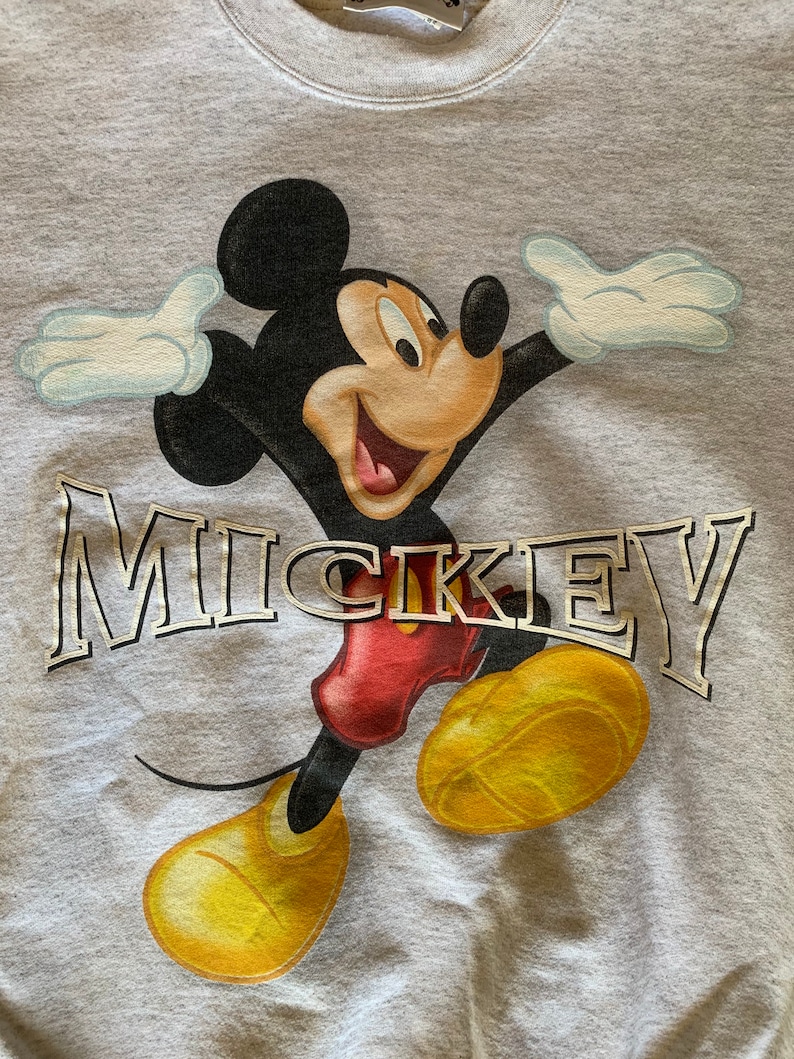 May include: A gray sweatshirt with a cartoon image of Mickey Mouse in a red and yellow outfit. The word "MICKEY" is printed in gold letters on the front of the sweatshirt.