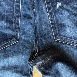 May include: Dark blue denim jeans featuring two back pockets with visible stitching. The right back pocket displays a small distressed tear, and the fabric shows a faded wash pattern throughout the material with reinforced seams at the crotch.