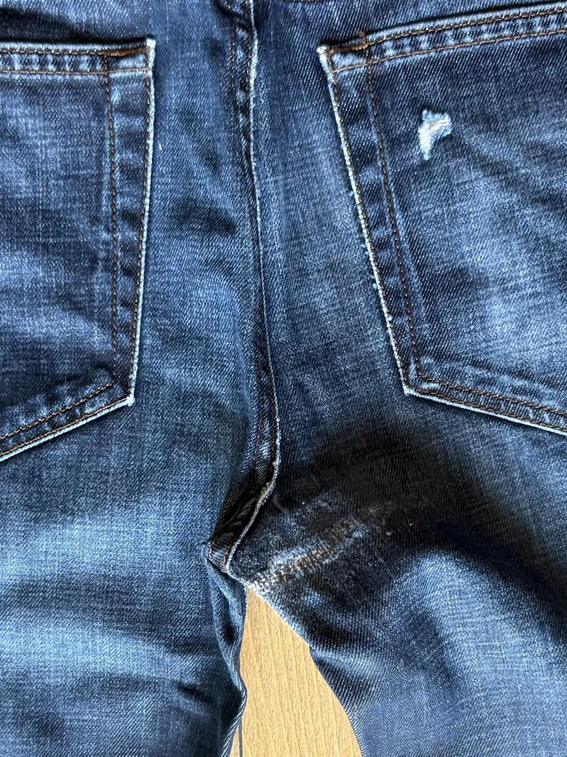May include: Dark blue denim jeans featuring two back pockets with visible stitching. The right back pocket displays a small distressed tear, and the fabric shows a faded wash pattern throughout the material with reinforced seams at the crotch.