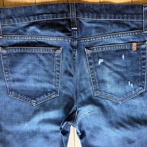 May include: A pair of dark blue denim jeans featuring a classic five-pocket design with a back view. The fabric displays a faded wash with subtle distressed details, including small frayed areas on the right back pocket and a small metal tag.