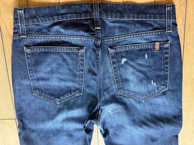 May include: A pair of dark blue denim jeans featuring a classic five-pocket design with a back view. The fabric displays a faded wash with subtle distressed details, including small frayed areas on the right back pocket and a small metal tag.