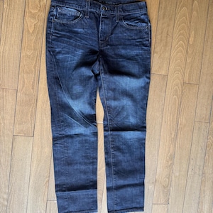 May include: A pair of dark wash denim jeans featuring a classic five-pocket design, a button and zipper fly closure, and belt loops. The fabric displays subtle whiskering and fading patterns across the thighs and hips with a straight leg cut.
