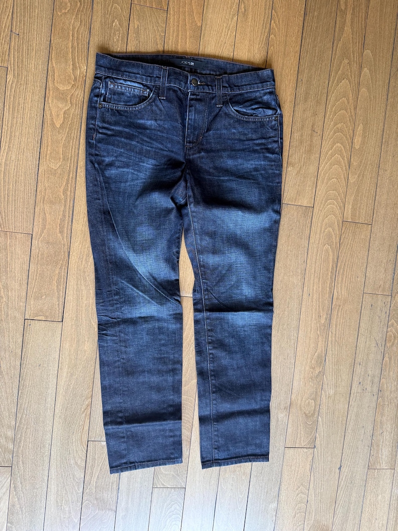 May include: A pair of dark wash denim jeans featuring a classic five-pocket design, a button and zipper fly closure, and belt loops. The fabric displays subtle whiskering and fading patterns across the thighs and hips with a straight leg cut.
