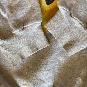 May include: Gray sweatshirt with a cartoon character design. The character is yellow and black and has a large head. The sweatshirt has ribbed cuffs.
