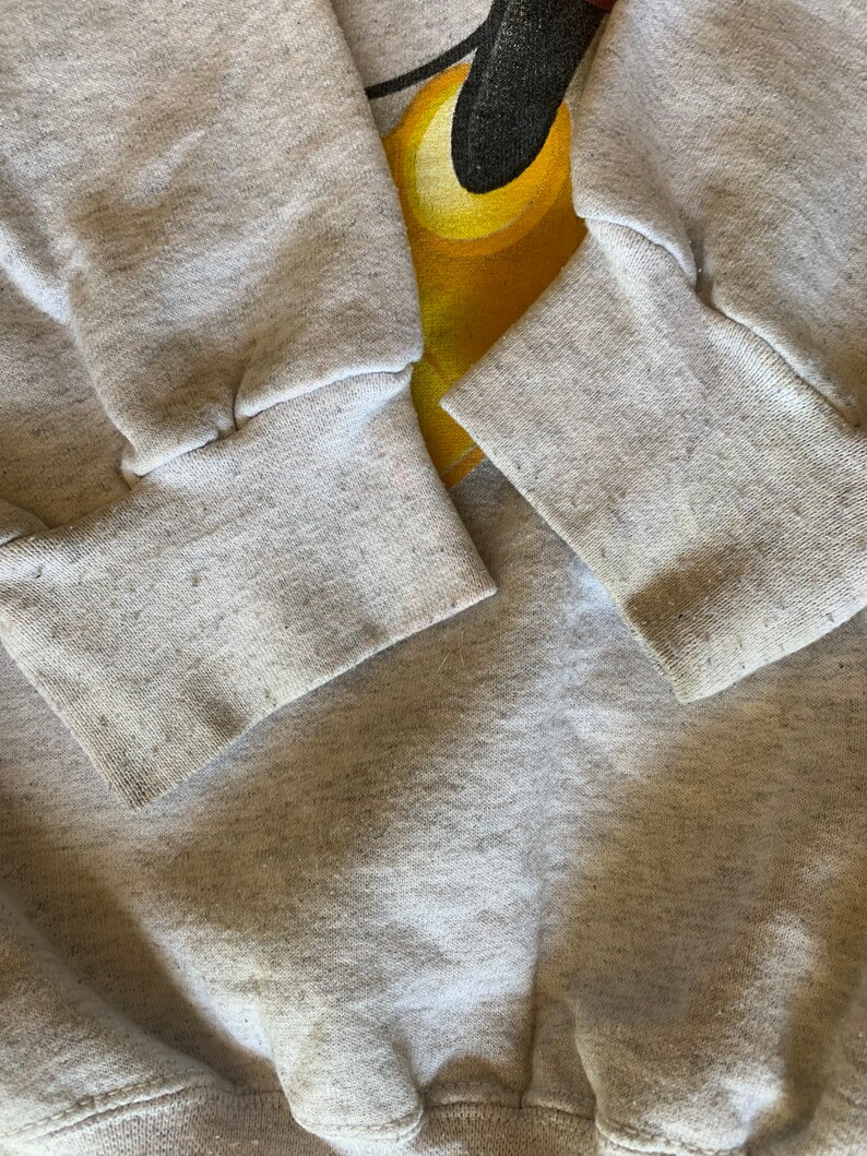 May include: Gray sweatshirt with a cartoon character design. The character is yellow and black and has a large head. The sweatshirt has ribbed cuffs.