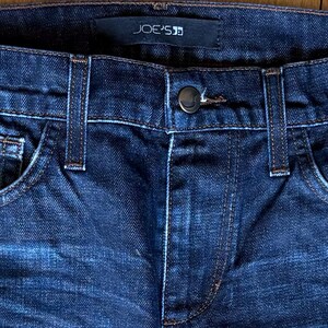 May include: Dark blue denim jeans featuring a classic five-pocket design with belt loops and a central metal button closure. A black fabric label with the white text JOE'S is stitched onto the interior waistband at the center back.