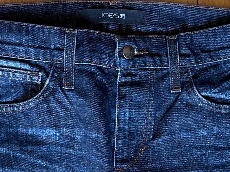 May include: Dark blue denim jeans featuring a classic five-pocket design with belt loops and a central metal button closure. A black fabric label with the white text JOE'S is stitched onto the interior waistband at the center back.