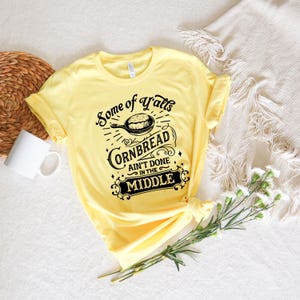 May include: Yellow t-shirt with a black graphic that says "Some of Y'all's Cornbread Ain't Done in the Middle". The graphic features a skillet with cornbread inside.