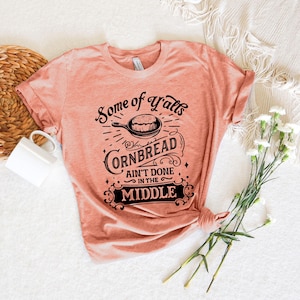 May include: A peach-colored t-shirt with a black graphic design that reads "Some of y'alls cornbread ain't done in the middle".