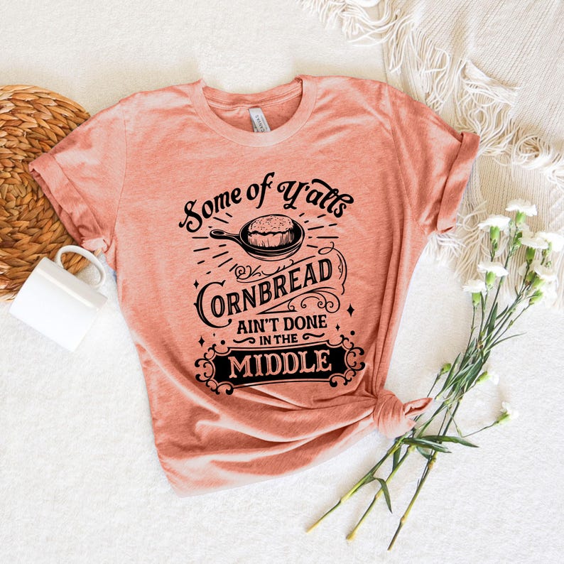 May include: A peach-colored t-shirt with a black graphic design that reads "Some of y'alls cornbread ain't done in the middle".