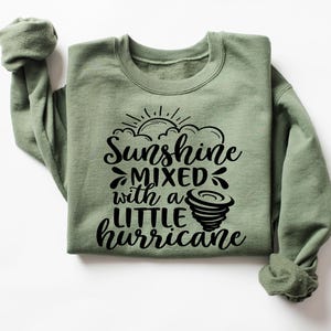 May include: Sage green sweatshirt with the text "Sunshine mixed with a little hurricane" in black. The design includes a sun, clouds, and a tornado graphic. The sweatshirt has a crew neck and long sleeves.