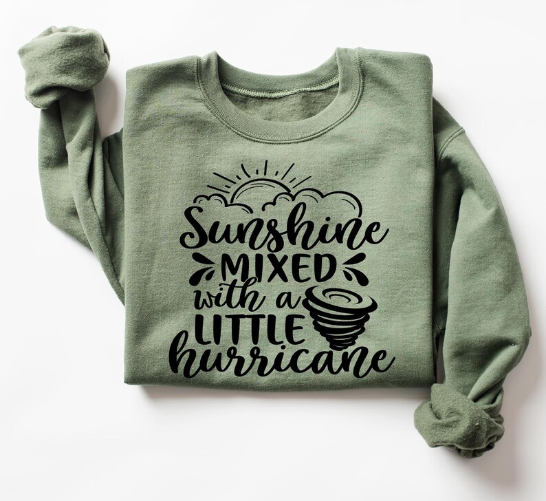 May include: Sage green sweatshirt with the text "Sunshine mixed with a little hurricane" in black. The design includes a sun, clouds, and a tornado graphic. The sweatshirt has a crew neck and long sleeves.