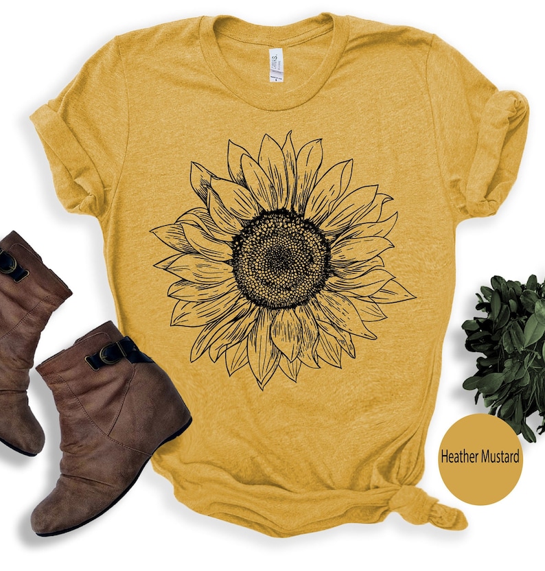 May include: A yellow t-shirt with a black line drawing of a sunflower. The shirt is made of a soft, comfortable material and has a relaxed fit. The text "Heather Mustard" is visible in the lower right corner of the image.