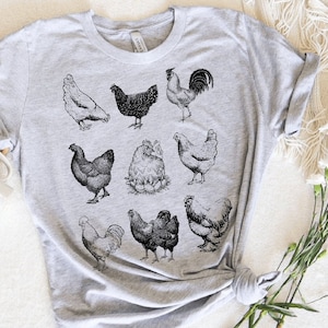 May include: A gray t-shirt with a black and white illustration of various chickens and roosters. The illustration includes a rooster with a large comb and wattles, a hen with a fluffy tail, and a group of chicks.