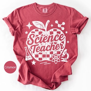 May include: Crimson-colored t-shirt with a white graphic. The graphic features an apple shape with a checkered pattern, the words "Science Teacher," and science-related icons. The shirt has rolled-up sleeves and a crew neck.