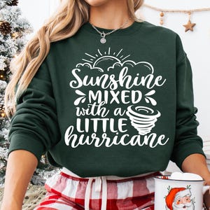 May include: Dark green sweatshirt with the text "Sunshine Mixed with a Little Hurricane" in white. The design includes a sun, clouds, and a tornado. The person is wearing red and white plaid pajama pants.