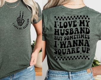 May include: A green t-shirt with a black skeleton hand making a peace sign on the front and the text "I love my husband" on the back. The back of the shirt also has the text "I love my husband but sometimes I wanna square up" with a checkered pattern.