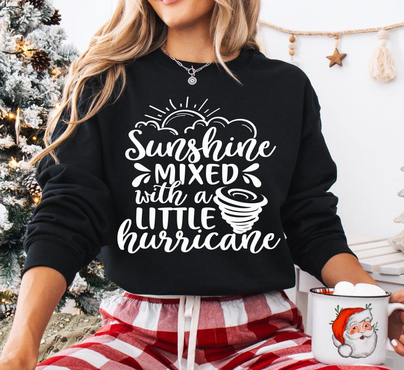 May include: A black sweatshirt with the text "Sunshine mixed with a little hurricane" in white. The design includes a sun, clouds, and a tornado. The person is wearing red and white plaid pajama pants and holding a Santa mug.
