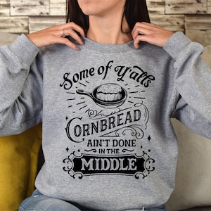 May include: Gray sweatshirt with a black graphic design that says "Some of y'all's cornbread ain't done in the middle". The graphic also includes a drawing of a pan of cornbread.