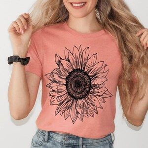 May include: A woman wearing a peach colored t-shirt with a black and white sunflower graphic print.