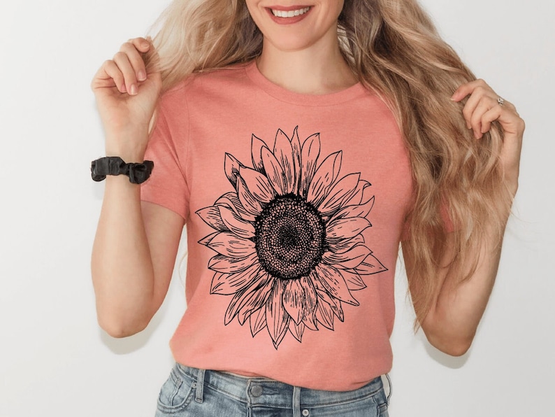 May include: A woman wearing a peach colored t-shirt with a black and white sunflower graphic print.