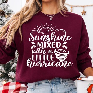 May include: Burgundy sweatshirt with the text "Sunshine Mixed with a Little Hurricane" in white script. The design includes a sun, clouds, and a tornado graphic. The sweatshirt is a crew neck style.