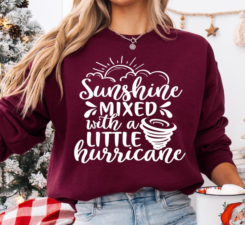 May include: Burgundy sweatshirt with the text "Sunshine Mixed with a Little Hurricane" in white script. The design includes a sun, clouds, and a tornado graphic. The sweatshirt is a crew neck style.
