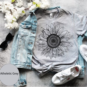 May include: A gray t-shirt with a black and white sunflower graphic. The shirt is laid out on a gray surface with a denim jacket, sunglasses, and white sneakers.