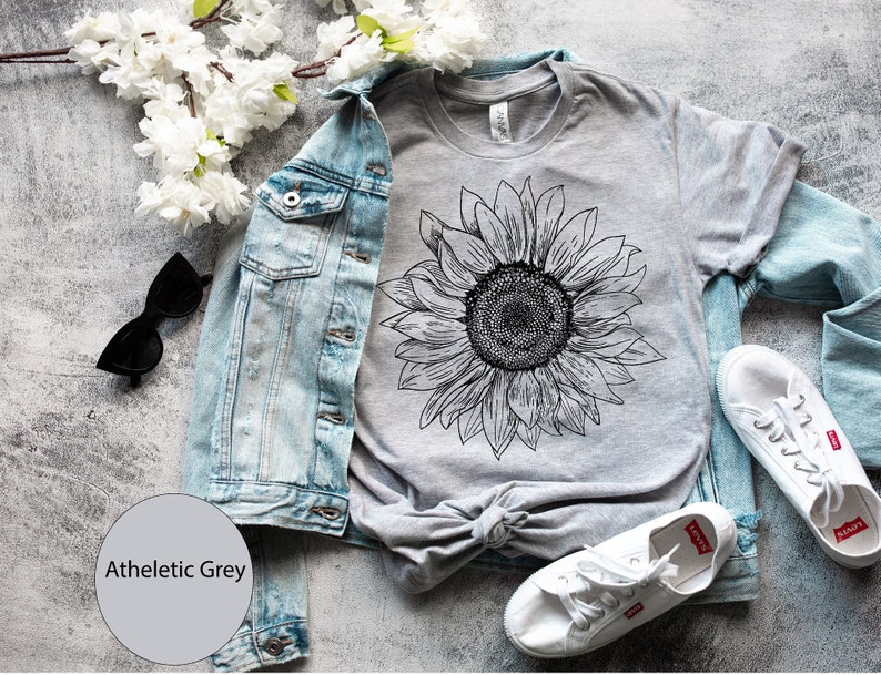 May include: A gray t-shirt with a black and white sunflower graphic. The shirt is laid out on a gray surface with a denim jacket, sunglasses, and white sneakers.