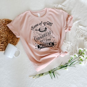 May include: A pink t-shirt with a black graphic that says "Some of y'all's cornbread ain't done in the middle". The graphic features a skillet with cornbread inside.