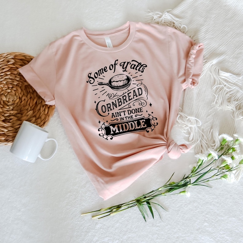 May include: A pink t-shirt with a black graphic that says "Some of y'all's cornbread ain't done in the middle". The graphic features a skillet with cornbread inside.