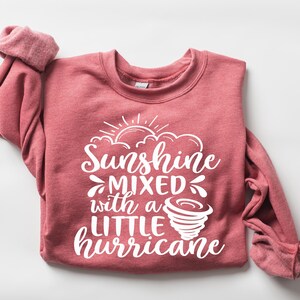 May include: A red-pink sweatshirt with the text "Sunshine mixed with a little hurricane" in white. The design includes a sun, clouds, and a tornado graphic. The long-sleeved top is folded.