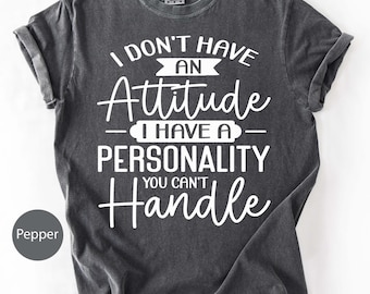 May include: Dark grey t-shirt with the text "I DON'T HAVE AN Attitude I HAVE A PERSONALITY YOU CAN'T Handle" in white script. The shirt has short sleeves and a crew neck. The color is labeled "Pepper".