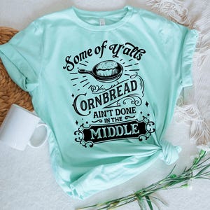 May include: A light blue t-shirt with a black graphic that says "Some of Y'alls Cornbread Ain't Done in the Middle". The graphic features a skillet with cornbread inside.