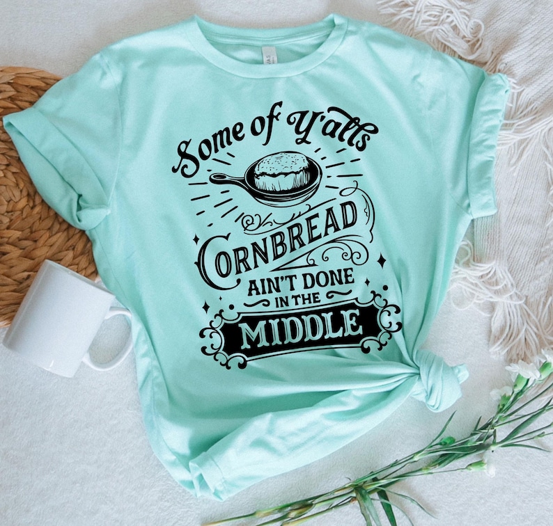 May include: A light blue t-shirt with a black graphic that says "Some of Y'alls Cornbread Ain't Done in the Middle". The graphic features a skillet with cornbread inside.