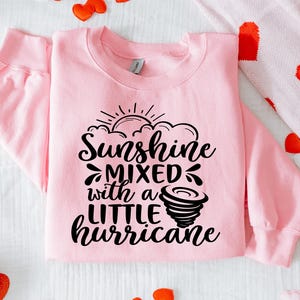 May include: A light pink sweatshirt with the text "Sunshine mixed with a little hurricane" in black script. The design includes a sun, clouds, and a tornado graphic. The sweatshirt has long sleeves and a crew neck.