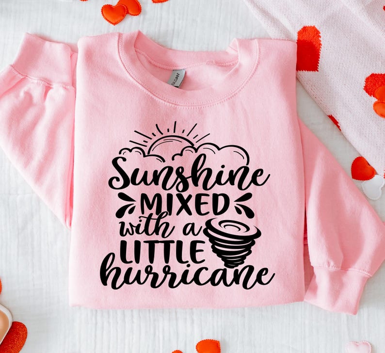 May include: A light pink sweatshirt with the text "Sunshine mixed with a little hurricane" in black script. The design includes a sun, clouds, and a tornado graphic. The sweatshirt has long sleeves and a crew neck.